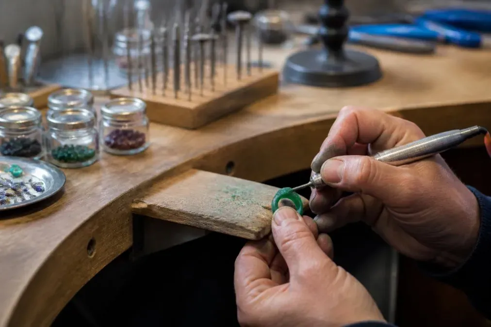Plan rapproché des mains d'un artisan joaillier travaillant une maquette en cire sur un établi d'atelier, avec bocaux de pierres précieuses et outils de sertissage en arrière-plan flou