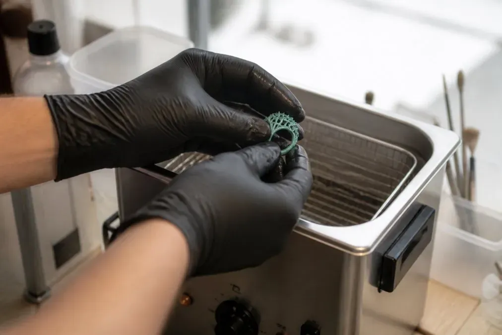 Mains gantées nettoyant des prototypes de bijoux en résine dans un bac d'alcool isopropylique en atelier.