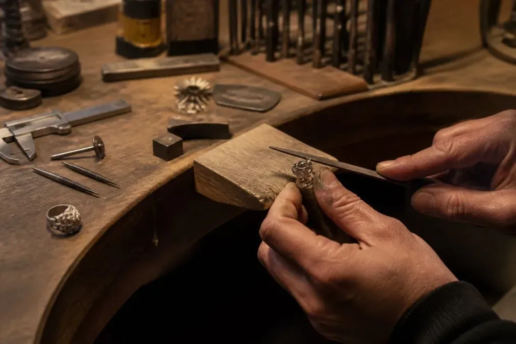 Artisan's hands working on a jewelry prototype with metal tools, files, and precious metal pieces on a wooden workbench under warm studio lighting