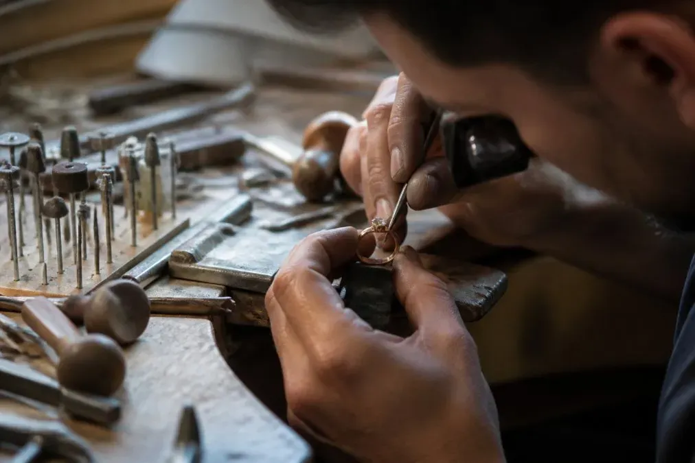 Artisan bijoutier travaillant à l'établi, mains en gros plan pendant le sertissage d'une pierre précieuse dans une bague en or, outils de joaillerie traditionnels visibles sous l'éclairage naturel