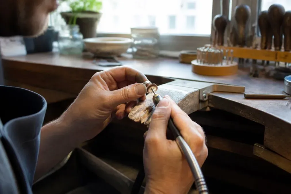 Gros plan des mains d'un artisan joaillier polissant une bague en argent à l'établi, outil rotatif en main, atelier lumineux avec lumière naturelle
